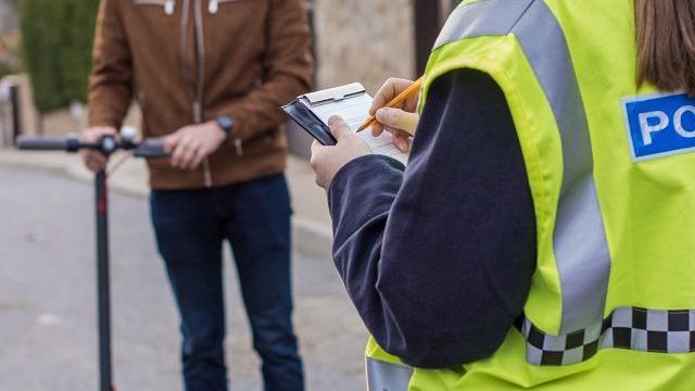 A person in a hi-vis police jacket taking notes with a dismounted e-scooter rider looking on