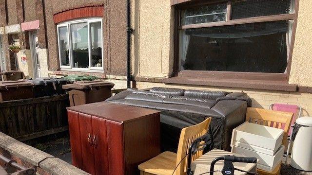 A sofa, cabinet, chairs and other furniture sit outside a property in a front garden