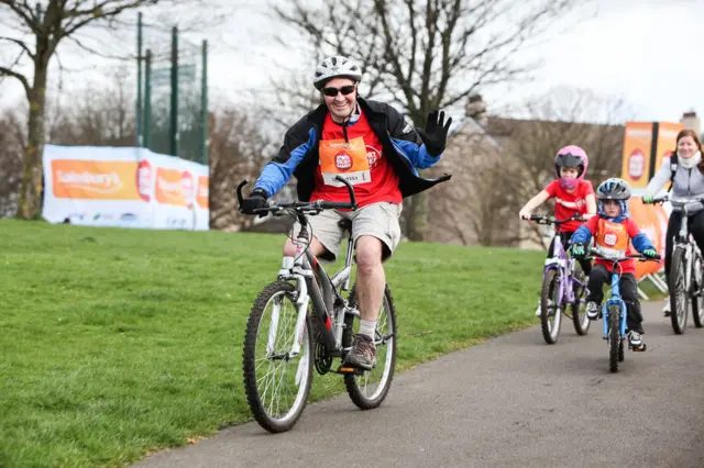 Man on his bike waving at camera followed by 2 children on bikes and a lady