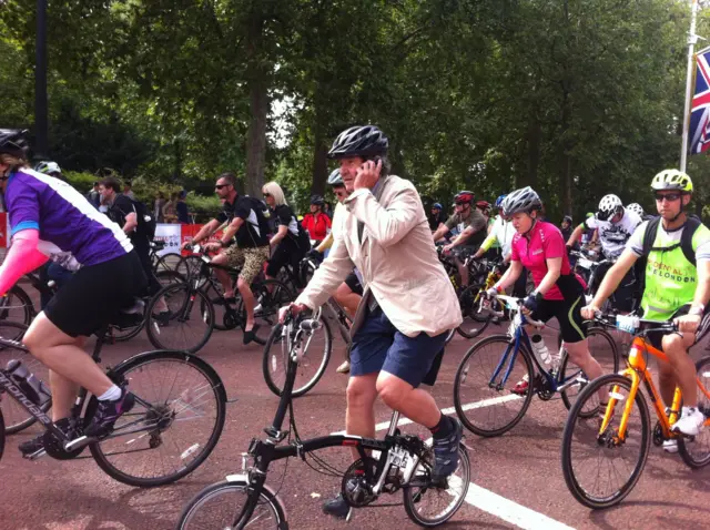 Many people on cycle bikes all sporty except a man on his fold up bike on the phone with a smart jacket on