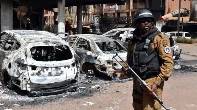 hotel charred with burnt out cars outside and a soldier with gun