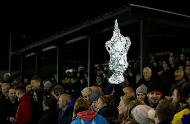 A fan holds up a replica cup wrapped in foil during the FA Cup First Round match between Warrington Town and Exeter City