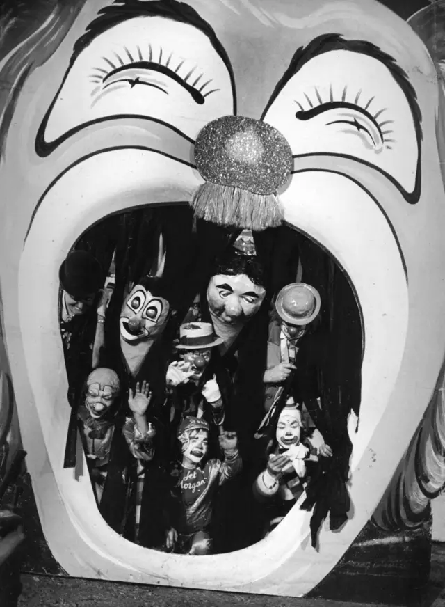 An old image of a theatre set with a clown's open mouth and several clowns waving and smiling.