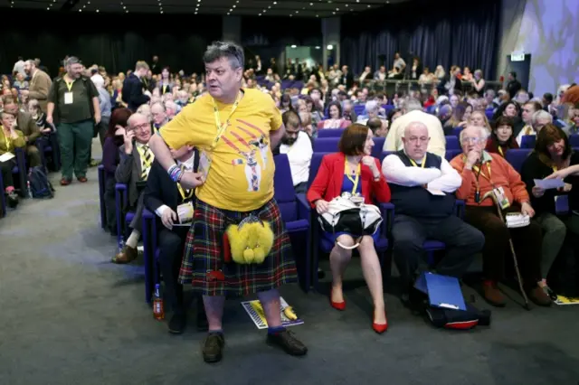 Man in kilt and yellow t-shirt standing by seated delegates