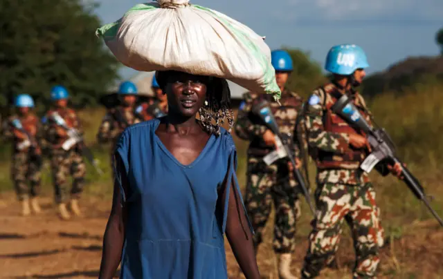 Woman carries sack on her head with soldiers in the background