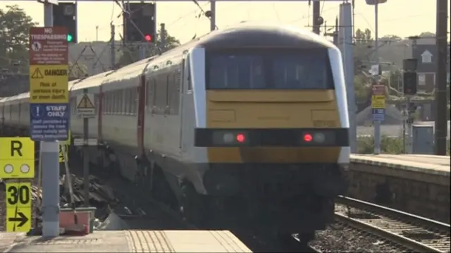 Train leaving the station, with overhead cables and rail signs warning of dangers of trespassing