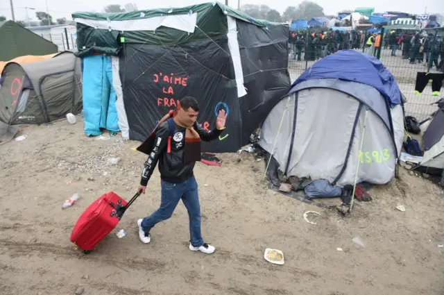 A man waving while leaving the "Jungle" camp with a suitcase