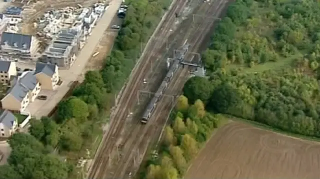 Aerial view of train on track, with housing to one side and farmland to the other