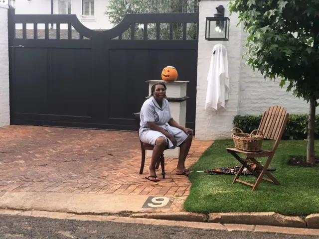 Woman sits outside gates of property with a wicker basket of treats on a chair