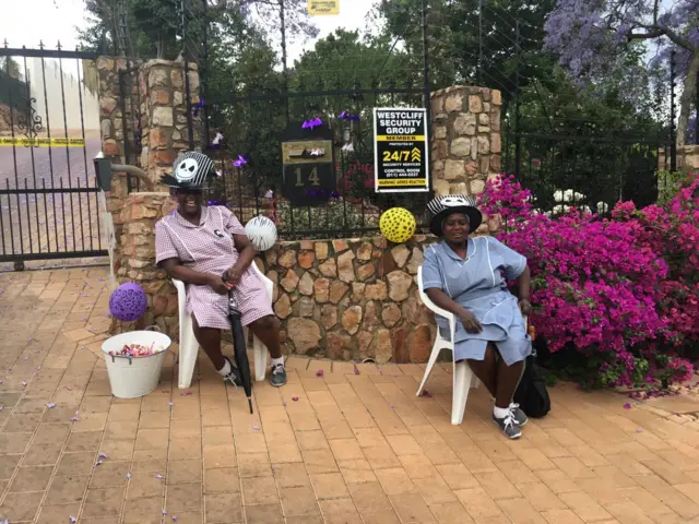 Two women in domestic uniforms sit outside a gated house with a bucket of sweets