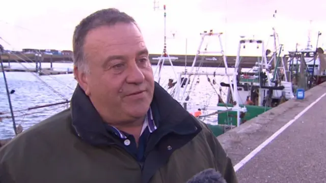 Paul Lines in brown coat, with fishing boats behind him