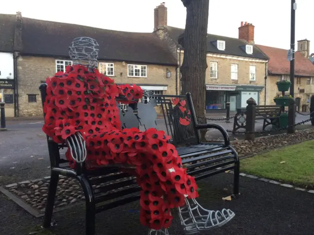 A sculpture of a man covered entirely in poppies sitting on a park bench in Higham Ferrers.
