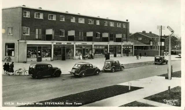 A black and white photo of Stevenage in the early fifties showing a row of shops and passing cars.