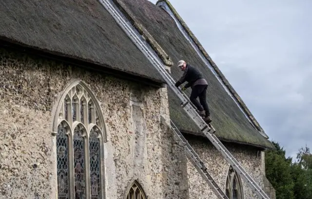 A workman on a ladder, leaning against the thatched roof of Caston church