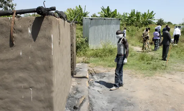 man collecting bodies to bury in a mass grave approaches a burned hut containing charred corpses