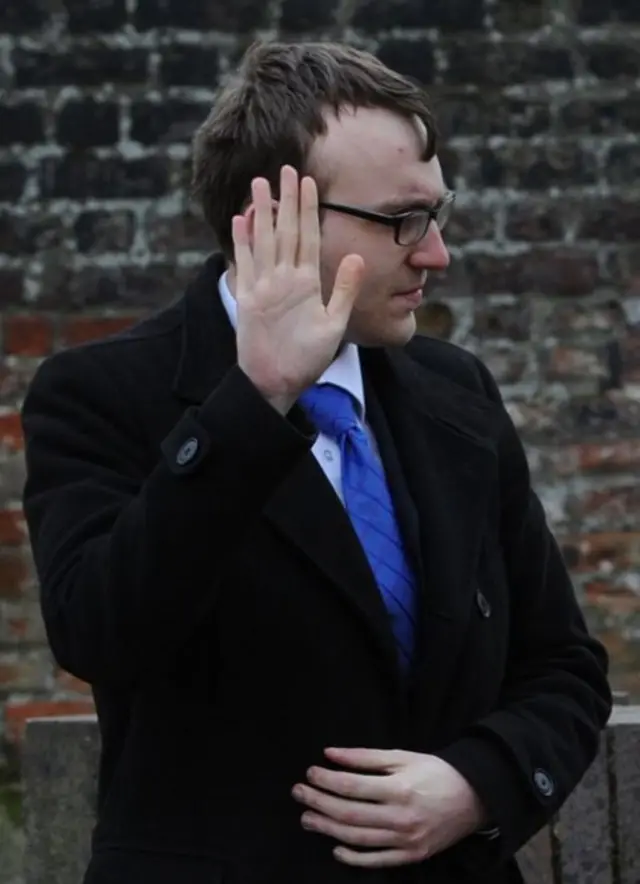 Luke Mallaband, wearing dark rimmed glasses, in a dark overcoat with bright blue tie, holding his right hand up to shield his face