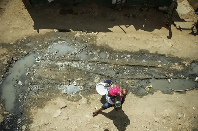 A stream of polluted water runs through as a resident from Zandspruit carries an empty bucket to be filled from one of the few communal water taps available to the thousands of people living in the Zandspruit informal settlement on March 11, 2015 in Johannesburg, South Africa