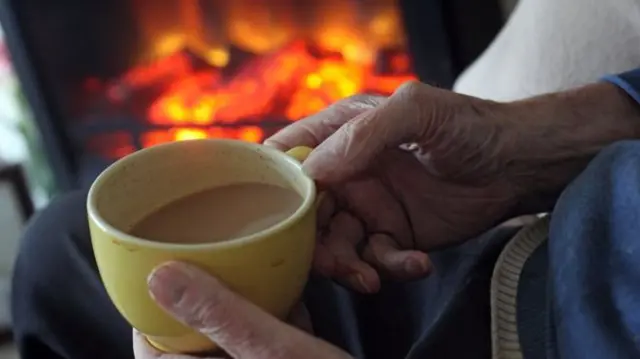 Elderly hand holding a cup of tea beside a coal fire