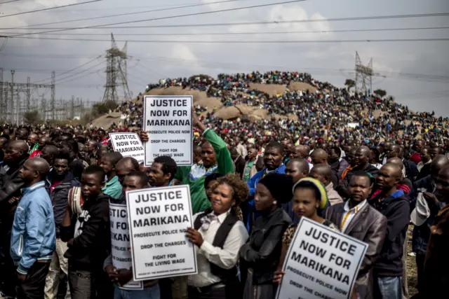 eople hold placards reading 'Justice now for Marikana, Prosecute police now, Drop the charges, Fight for the living wage' as people attend on August 16, 2014 in Marikana, .