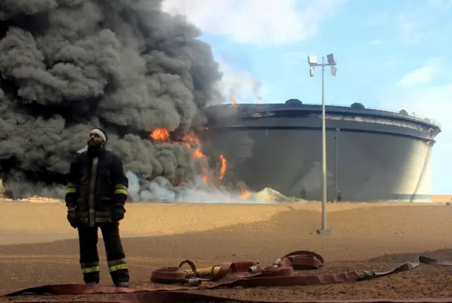 A Libyan fireman stands in front of smoke and flames rising from an oil storage tank