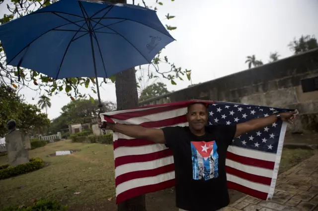 A Cuban man wearing a tee-shirt with a Cuban flag emblem and carrying the Stars and Stripes waits in the rain to wave to President Barack Obama"s convoy as it arrives in Old Havana, Cuba