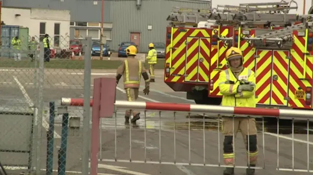 Scene at Norwich Airport, with fire crew by barrier, and hangars in background