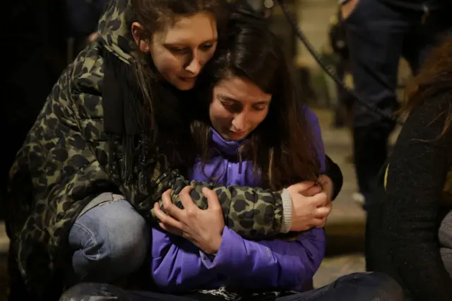 Young women hold each other at a makeshift memorial in front of the stock exchange at the Place de la Bourse