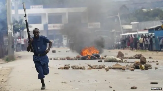 Burundi police officer runs through streets of Bujumbura with a gun, with fire burning in background