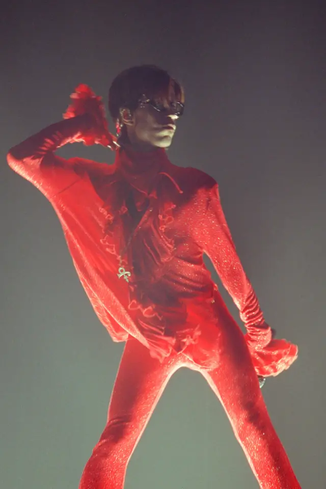 Prince onstage in a red frilled jumpsuit, looking assertive and camp, pumping his hair up