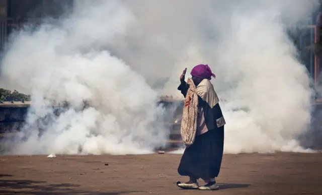 An elderly woman caught up in the clashes holds her hands in the air as a riot policeman approach amidst clouds of tear gas, during a protest in downtown Nairobi, Kenya Monday, May 16, 2016.