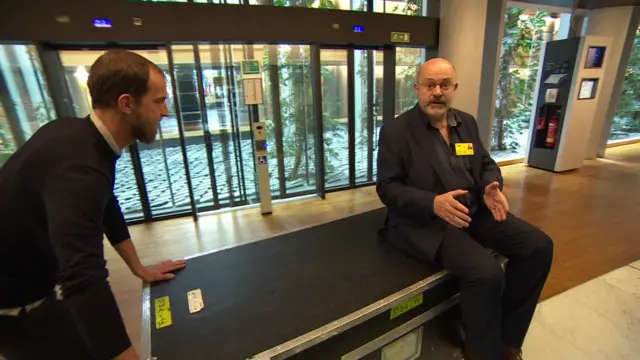 John Sweeney sits on a box being pushed through the European Parliament