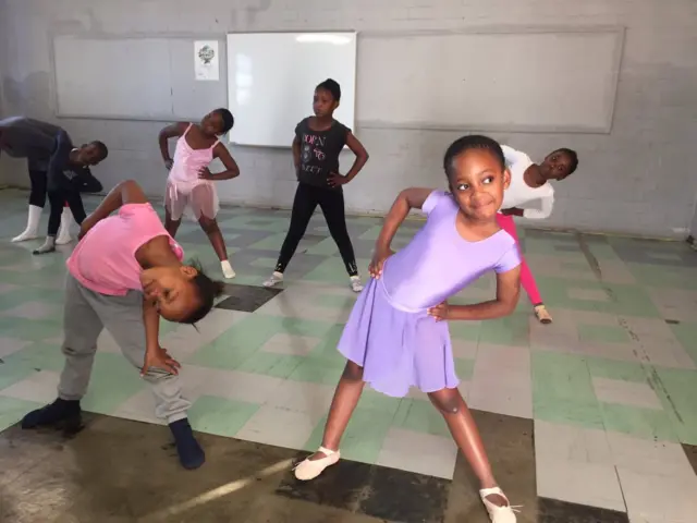 Kids strike a range of poses in ballet class. One girl in purple leotard grins broadly.