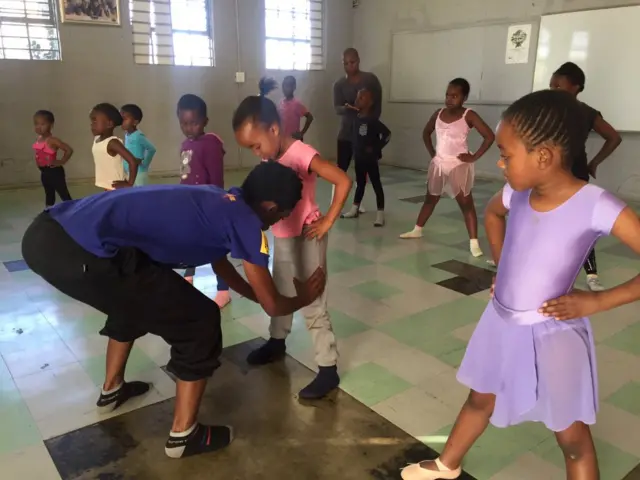 Children stand with legs apart as one young boy is shown the correct ballet stance by a teacher