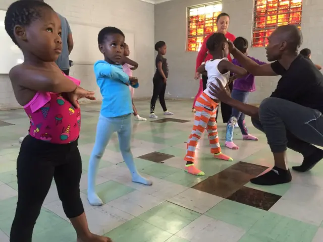 Children stand with legs apart but right arms across their chest in ballet class