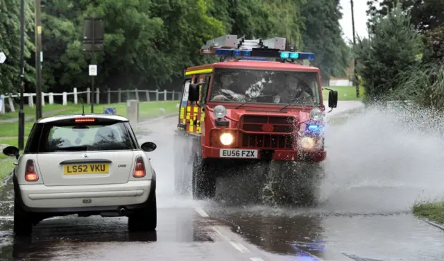 A driver gives way to an emergency vehicle as it drives through a flooded road in East Hanningfield, Essex