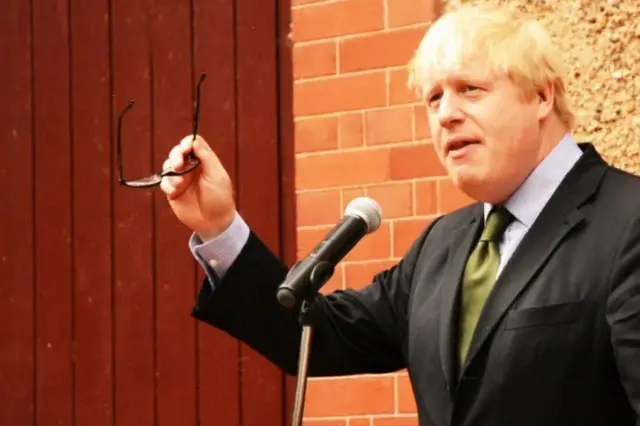 Boris Johnson standing by a microphone and holding his glasses