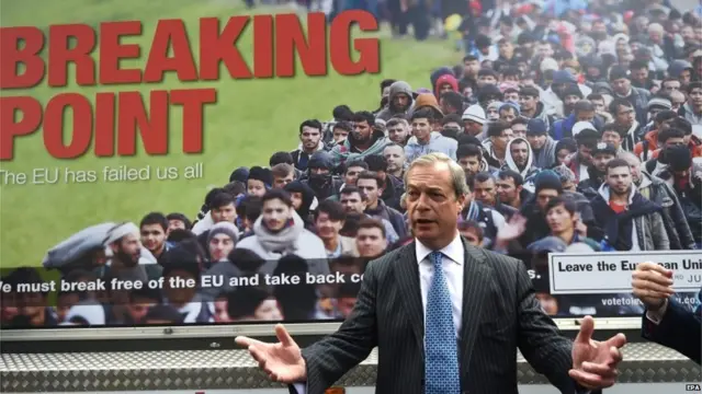 MEP Nigel Farage standing in front of a poster showing a long queue of migrants.