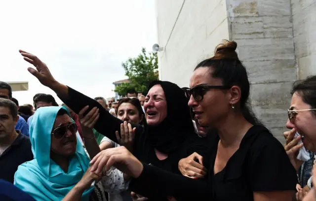 woman in black mourning clothes crying and reaching out, held back by others