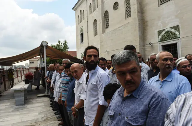 men line the side of a street, four-deep, looking in the same direction