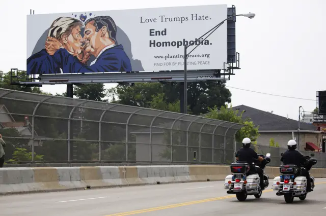 Bicyclists ride past a billboard depicting a kiss between Donald Trump and Ted Cruz on the first day of the Republican National Convention.