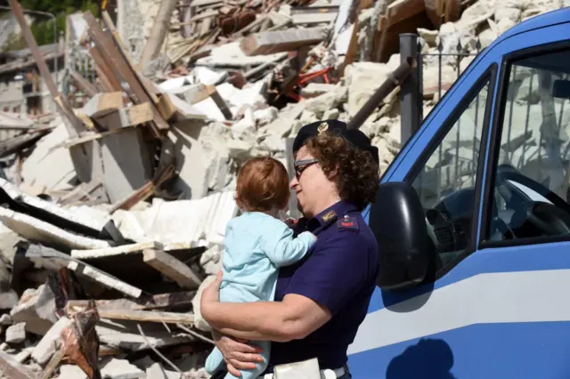 A police officer holding a child inspects collapsed and damaged houses in Pescara del Tronto, central Italy, 24 August 2016,