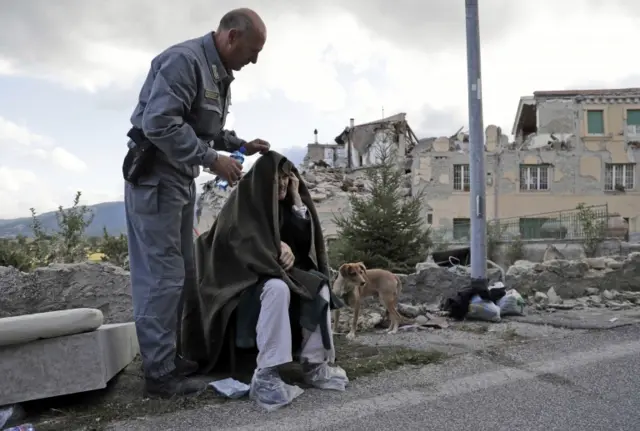 An elderly man is given assistance as collapsed buildings are seen in the background following the earthquake in Amatrice