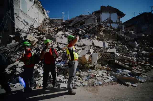 Rescuers standRescue workers stand next to destroyed buildings in Amatrice, central Italy (25 August 2016)