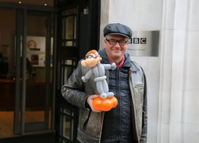 Charlie Higson, in leather jacket and cap, holding a balloon model, outside the entrance to BBC studios in London