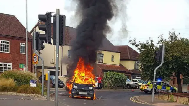 A police car on the road, to the right of the burning Smart car