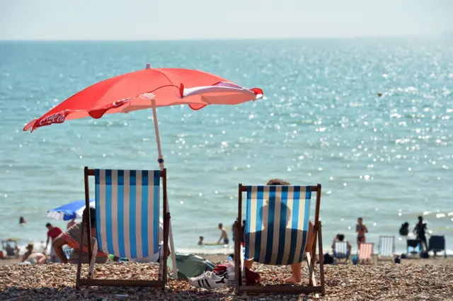 Two blue-striped deckchairs, on a pebbly beach, with a red umbrella and beach visitors