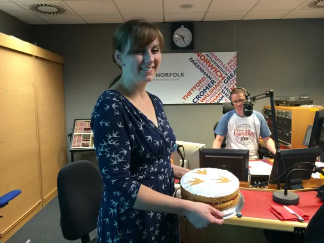 Kate Barmby, holding a sponge cake in radio studio, with Matthew Gudgin behind her at his radio desk