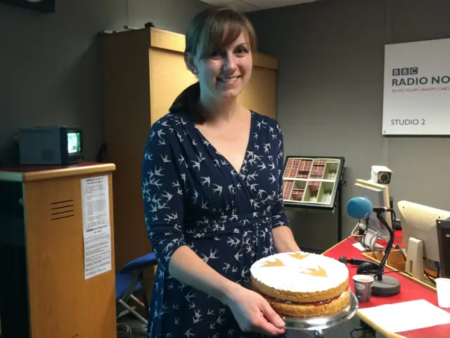 Kate Barmby, in blue patterned dress, holding a sponge cake in the radio studio