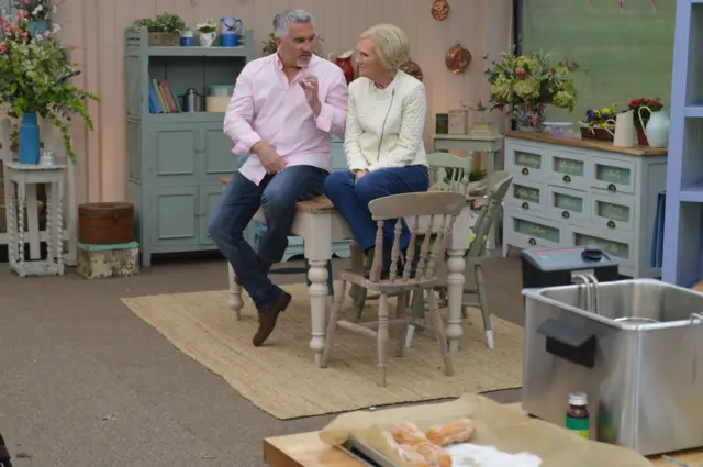 Paul Hollywood, in pink shirt and blue jeans, Mary Berry in cream jacket and blue trousers, sitting on a table in the Bake Off studio