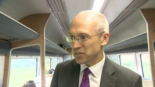 Jonathan Denby, in grey suit, white shirt and purple tie, standing in a train carriageway